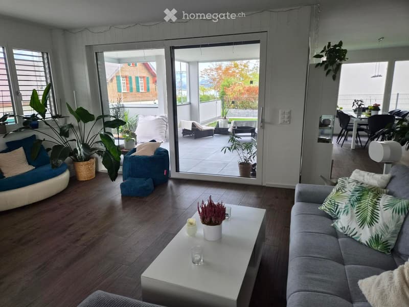 Modern living room with wooden flooring, large windows, and a sliding glass door leading to a balcony. Features a grey sectional sofa, white coffee table, and various potted plants.