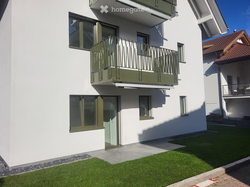 Modern two-story building with white exterior, green-framed windows, and a balcony with metal railing. The ground floor has a small lawn area and a paved pathway.