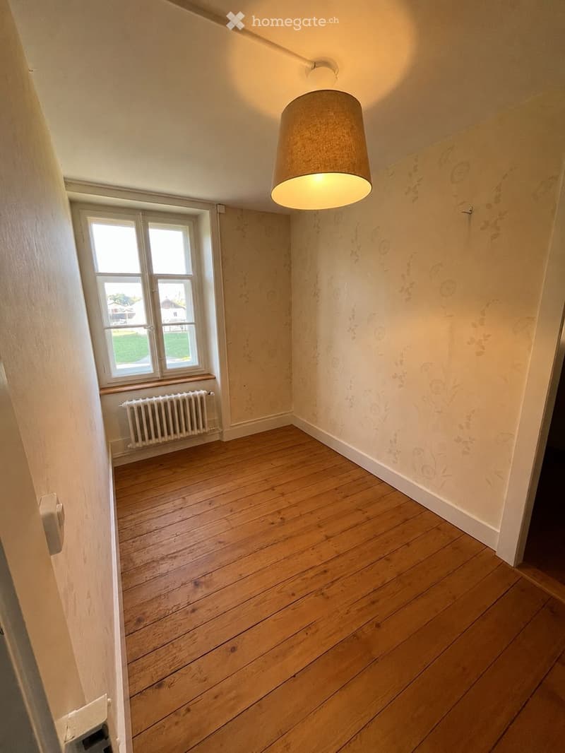 Empty room with wooden flooring, white walls, a window with a radiator below, and a hanging light fixture.