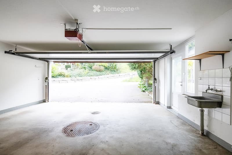 Empty garage with a drain in the floor, a sink on the wall, a shelf, and a door leading outside