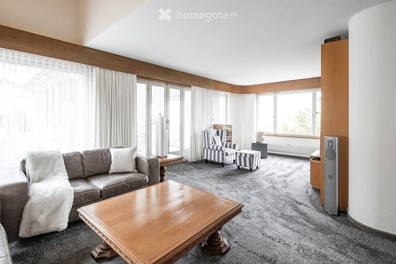 A cozy living room with a sofa, armchair, ottoman, center table, carpet, sliding glass doors, white curtains, and wooden ceiling trim.