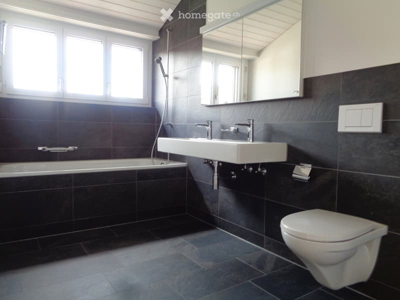 Modern bathroom with dark tiled walls and floor, featuring a bathtub, double sink, toilet, and large mirror. The room has large windows allowing natural light to enter.