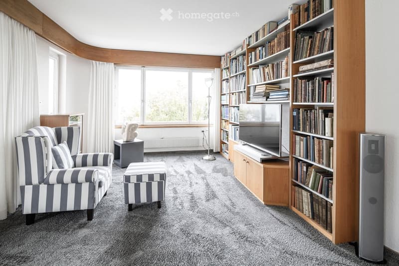 Cozy living room with grey and white striped sofa, matching ottoman, bookshelf filled with books, television, floor lamp, and decorative items.