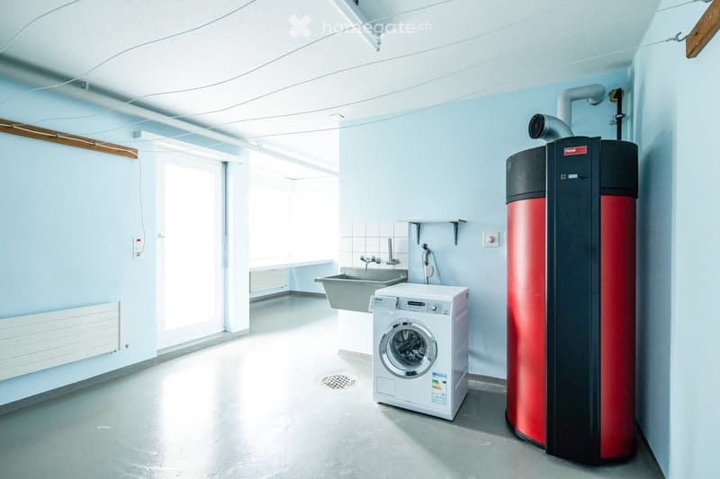 Bright, modern bathroom with a washing machine, sink, radiator, and water heater. Light blue walls and a large window provide natural light.