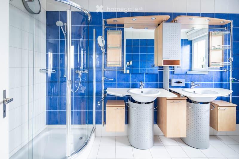 Modern bathroom with blue tiled walls, white tiled floor, double sink vanity with mirrored cabinets, and a glass-enclosed shower area.