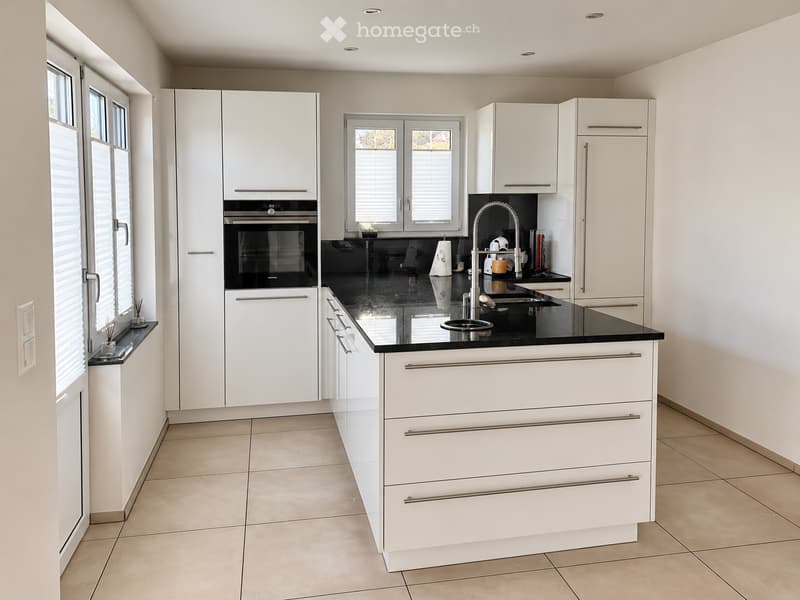 Modern kitchen with white cabinets, black countertop, stainless steel sink, and built-in oven. Features large windows and tiled flooring.