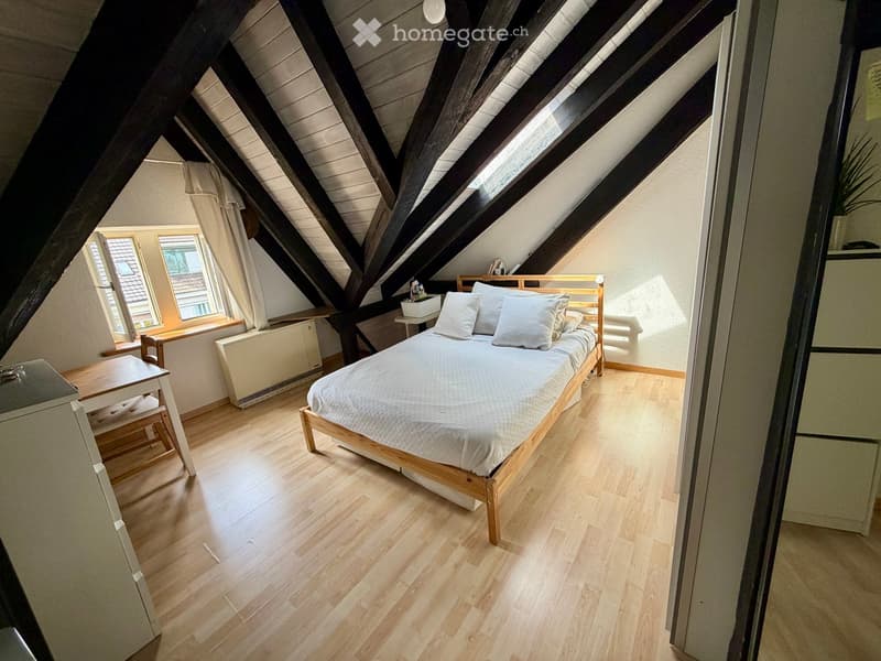 Cozy attic bedroom with wooden floors, slanted wooden ceiling beams, large windows, a single bed with white bedding, a small table, and a chair.