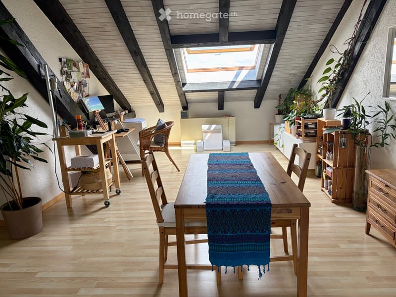 Bright attic room with wooden floors, slanted wooden ceiling beams, skylight window, dining table with chairs, desk with computer, and potted plants.