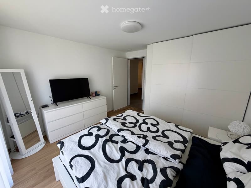 Modern bedroom with a large bed, white dresser, mirror, television, and nightstand. The room features light wooden flooring and white walls.
