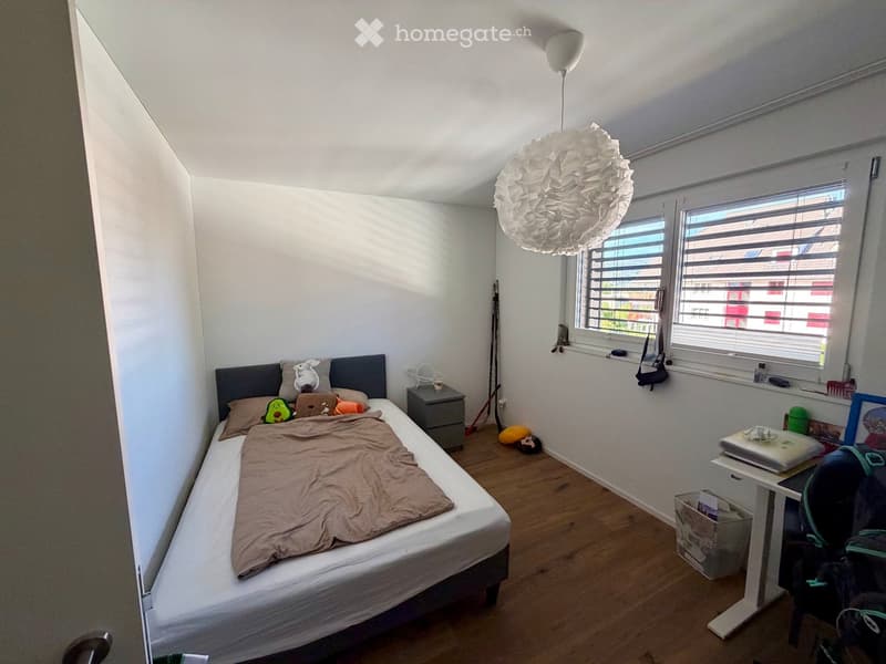 Cozy bedroom with a single bed, nightstand, laptop on a small table, and various personal items including toys and a backpack.