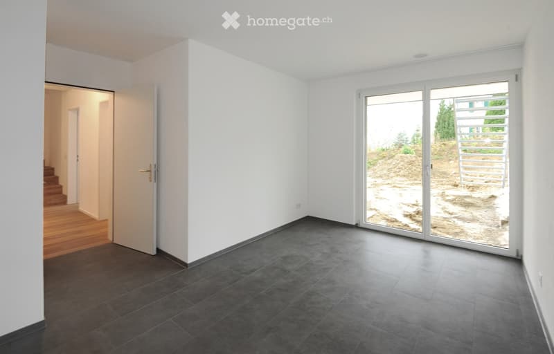Empty room with white walls, dark tiled floor, large sliding glass door leading outside, and an open doorway to another room with wooden flooring.