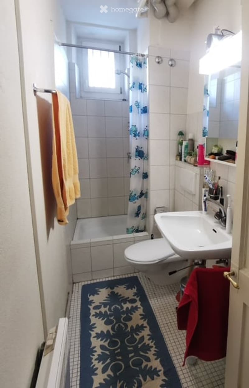A compact bathroom with white tiled walls and floor, featuring a bathtub with a shower curtain, a toilet, a sink with a mirror, and a towel rack with towels.