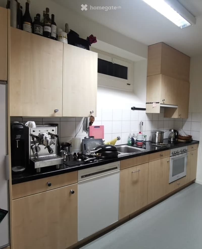 Modern kitchen with light wooden cabinets, black countertops, and various appliances including a coffee machine, stove, and oven. Bottles are displayed on top of the cabinets.