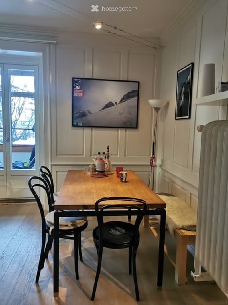 A small, cozy dining area with a wooden table, four black chairs, a teapot, cups, and a large TV mounted on the wall. The room has light-colored walls, wooden flooring, and a large window with a view of the outside.