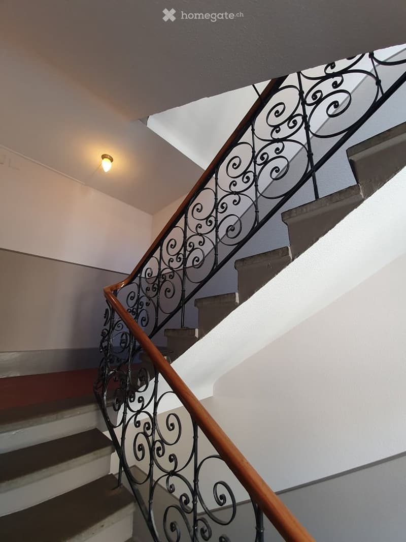 Staircase with wooden handrail and ornate metal balustrade, white walls, and a single ceiling light fixture.