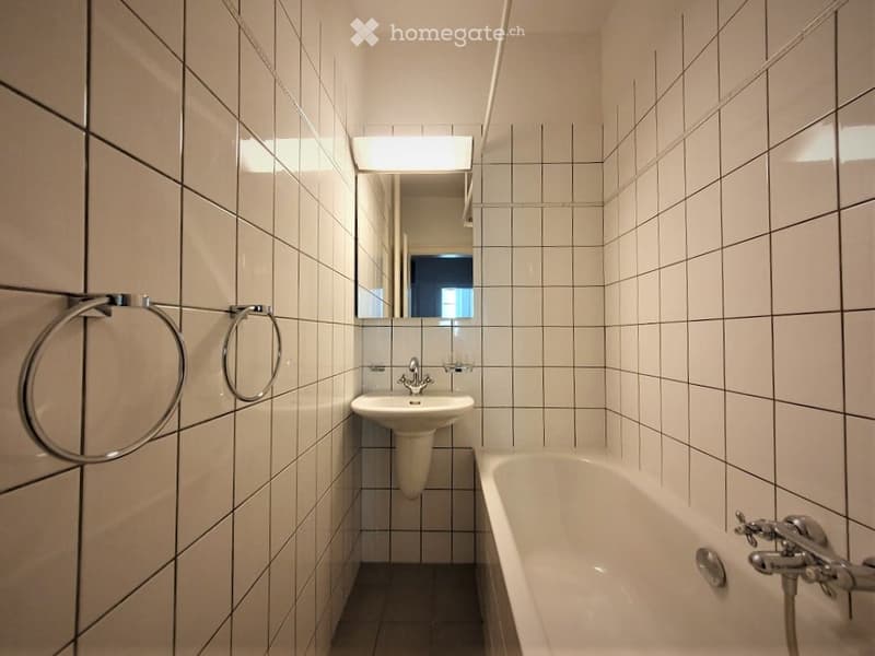Modern bathroom with white tiled walls and floor, featuring a sink, bathtub, mirror, and towel ring.