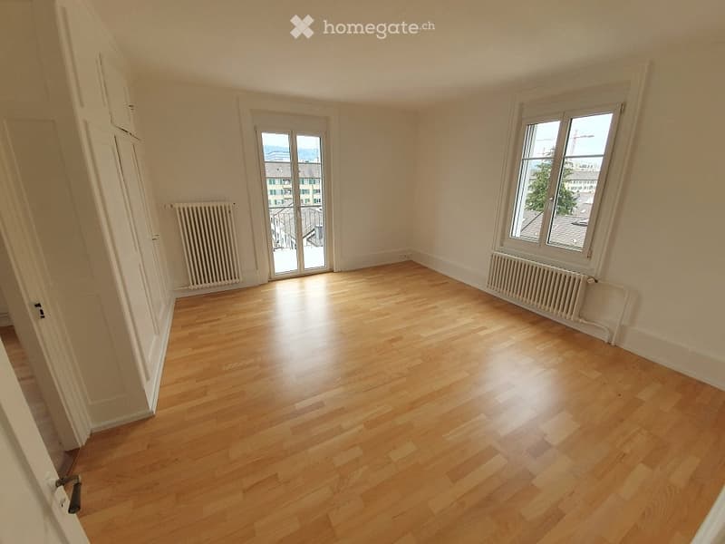 Empty room with wooden flooring, white walls, and large windows offering a view of the cityscape.