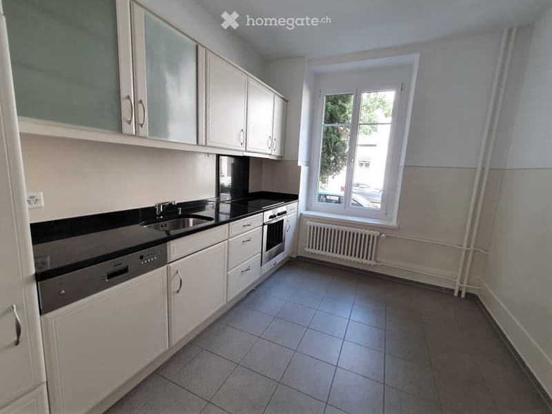 Modern kitchen with white cabinets, black countertops, and stainless steel appliances including a sink, oven, and dishwasher. A window with a view of greenery is visible.