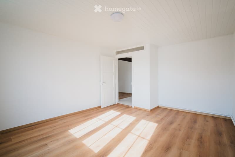 Empty room with white walls, wooden flooring, and a single open door leading to another space.