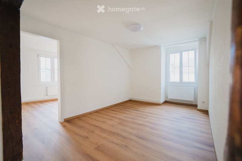 Empty room with wooden flooring, white walls, and large windows allowing natural light. The room features a slanted ceiling and a visible radiator under one window.