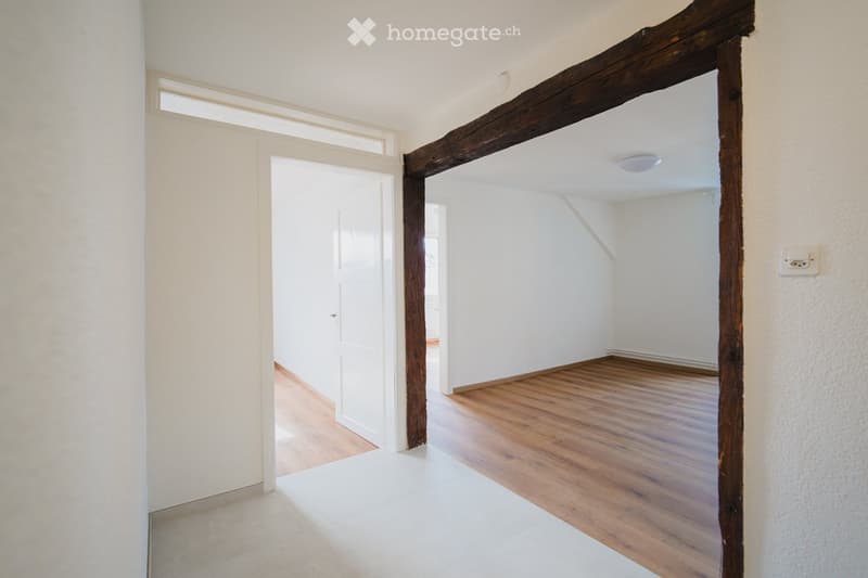Empty hallway with wooden floor, white walls, and exposed wooden beams. Features a white door and a small window allowing natural light to enter.