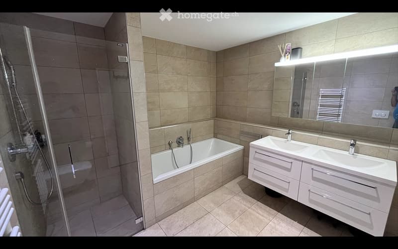 Modern bathroom with a bathtub, double sink, large mirror, and glass-enclosed shower. Beige tiles and white cabinetry.