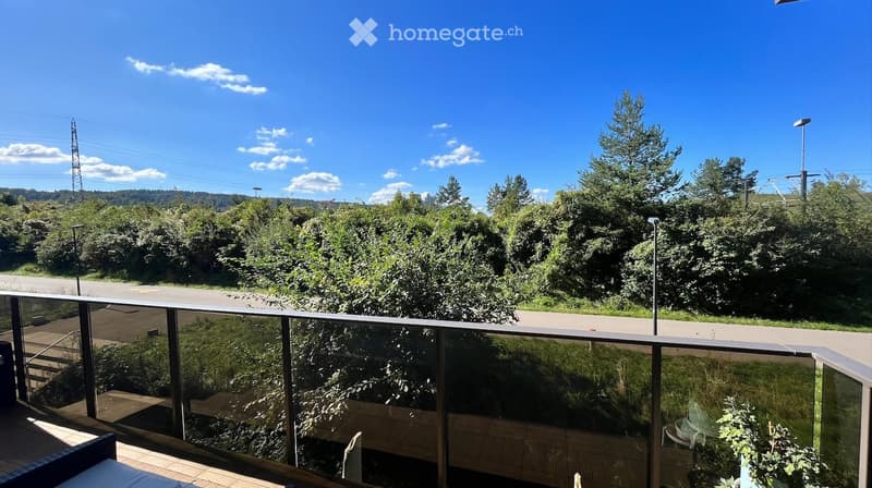 Balcony with glass railing, wooden decking, and outdoor furniture, overlooking a green, tree-filled landscape with a road below.