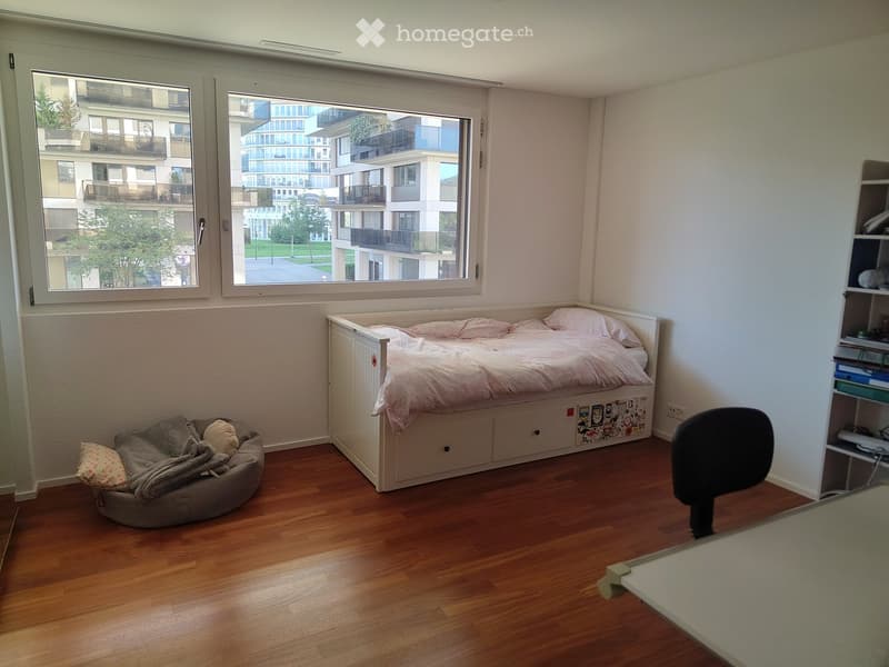 Cozy bedroom with a wooden floor, a single bed with pink bedding, a dog bed, a chair, and a shelf with various items.
