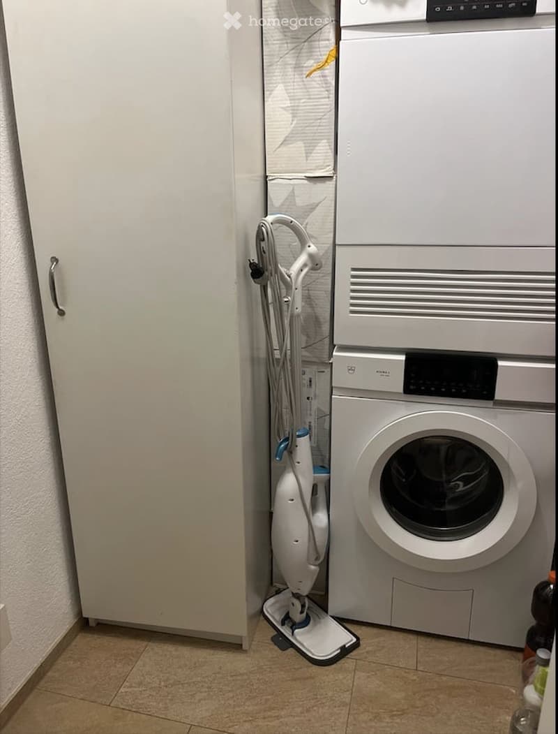 A compact bathroom with a washing machine, a white wardrobe, and a vacuum cleaner placed next to it.