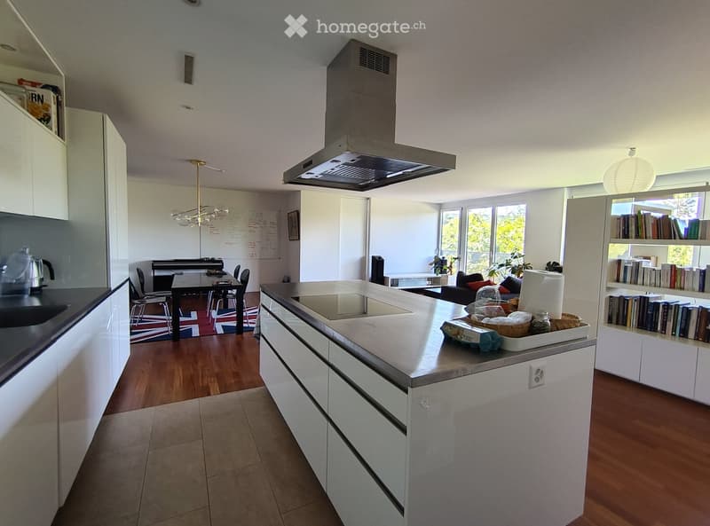 Open-plan kitchen and living area with white cabinets, a central island, a dining table with chairs, a sofa, and a bookshelf filled with books. The space features wooden flooring and large windows letting in natural light.