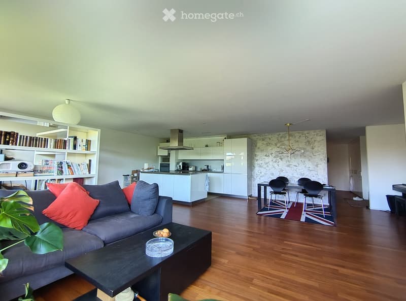 Modern open-plan living and dining area with a grey sectional sofa, black coffee table, bookshelf, white kitchen with cabinets, and a dining table with chairs.