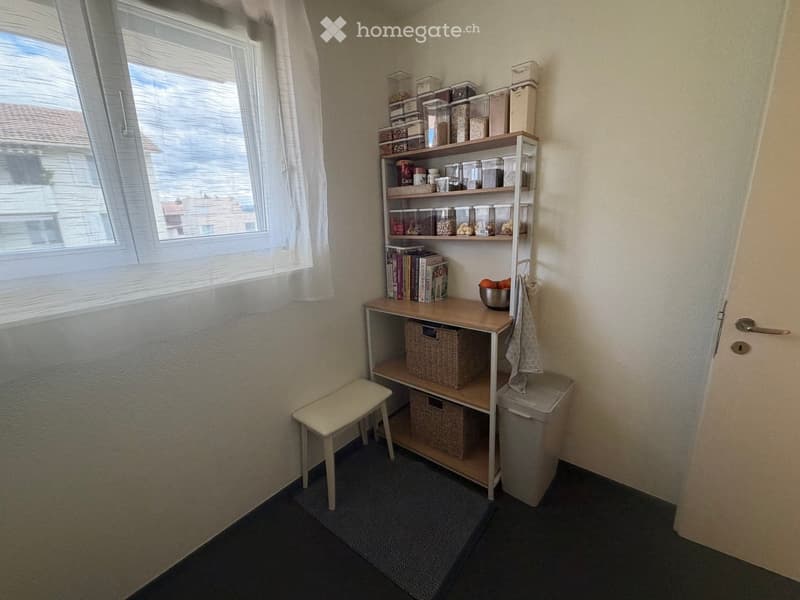 A small, well-lit room with a window, a stool, a table with shelves holding books and a bowl, and baskets underneath.