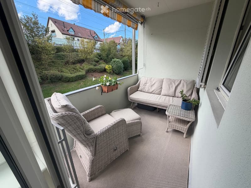 Balcony with a sofa, wicker chair, small table, and potted plants, overlooking a green garden.