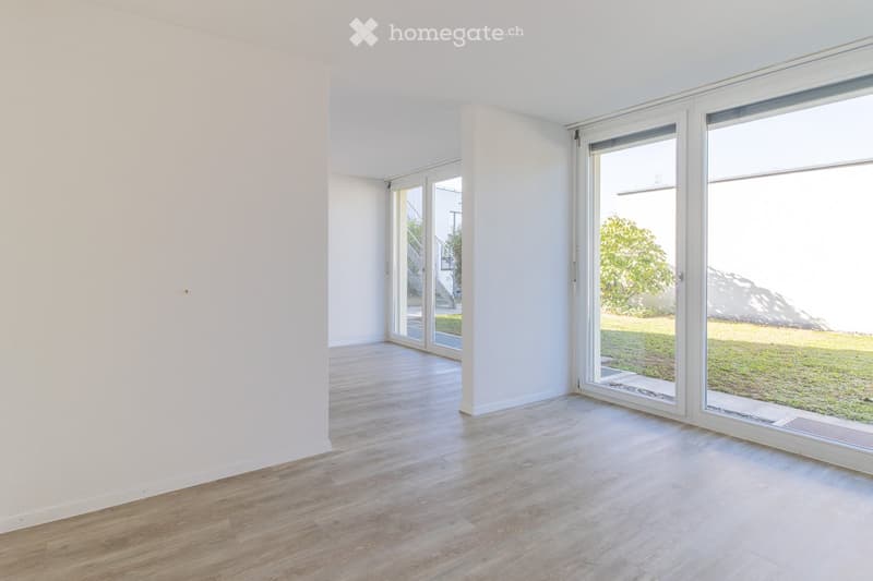 Empty room with light wooden flooring and white walls, featuring large glass sliding doors that open to a grassy outdoor area.