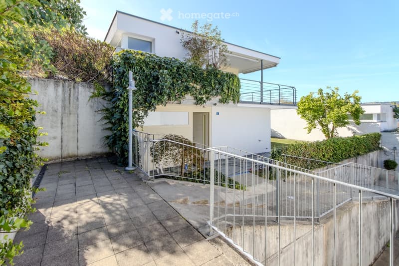 Modern two-story house with white exterior, raised ground floor, and balcony on the upper level. The entrance features a metal railing and steps leading up to the main door.