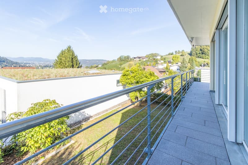Balcony with metal railing, tiled floor, overlooking a green landscape with trees and distant hills.