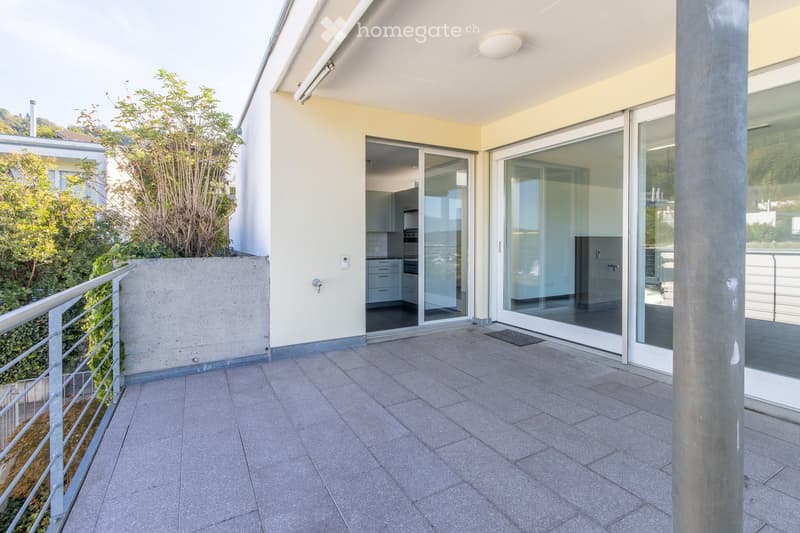 Modern balcony with tiled flooring, large sliding glass doors leading to a kitchen area, metal railing, and a view of greenery.