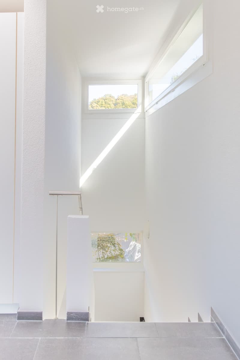 Modern, minimalist hallway with white walls, grey tiled floor, and large windows allowing natural light to flood the space. The hallway features sleek, contemporary design elements and a clean, uncluttered aesthetic.