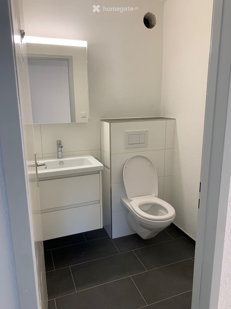 Modern bathroom with white walls, black tiled floor, sink with chrome faucet, mirror, and toilet with open lid.