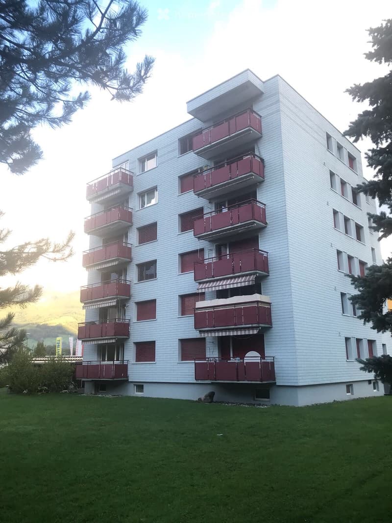 A modern multi-story apartment building with white siding and red balconies, surrounded by green grass and trees.