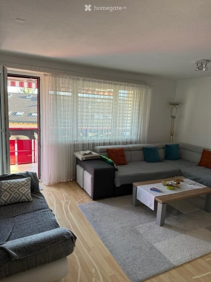 Modern living room with grey sectional sofa, coffee table, and large windows with sheer curtains. The room features a wooden floor and a grey rug.