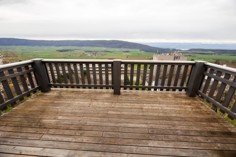 Wooden balcony with grey railing, overlooking a scenic countryside with fields, hills, and distant mountains.