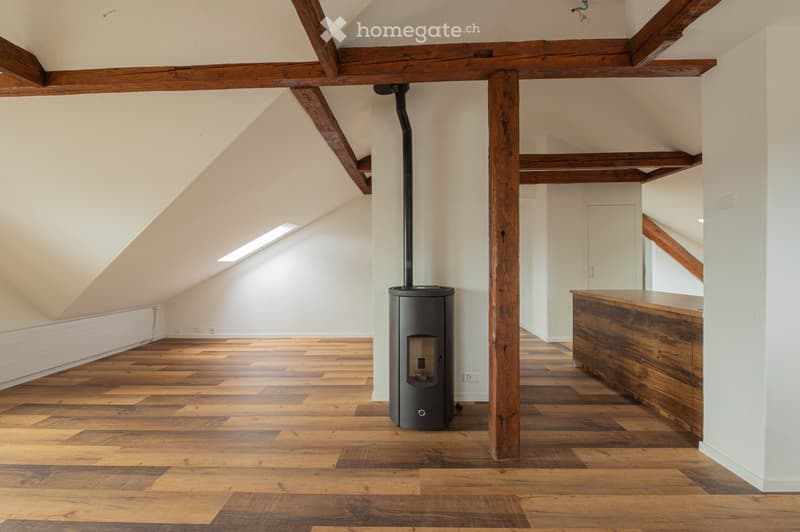 Open loft-style room with wooden beams, polished wooden floor, and a modern black wood-burning stove.