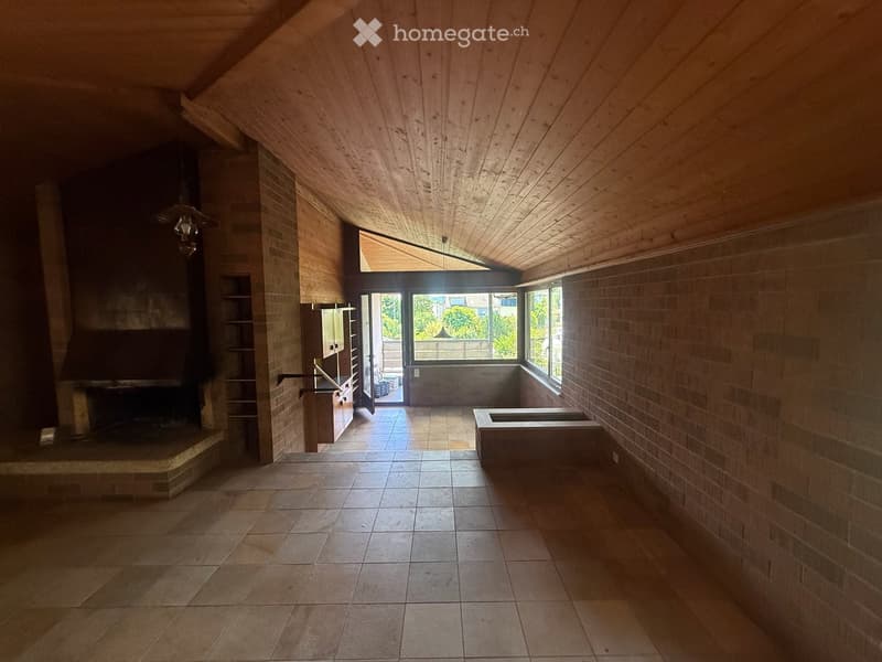 An unfurnished room with exposed wooden beams on the ceiling, brick walls, tiled floor, and a large fireplace. There is a doorway leading outside with a view of greenery.