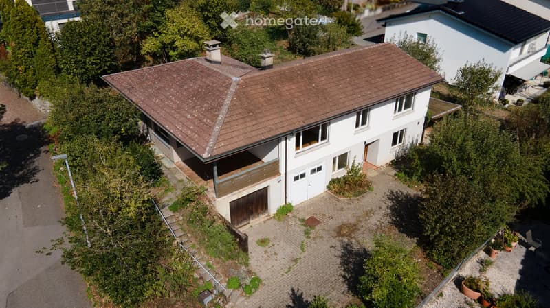 Aerial view of a two-story house with a brown tiled roof, white exterior, and a garage. The house features multiple windows and a small balcony area. The surrounding area includes a driveway, garden, and neighboring houses.