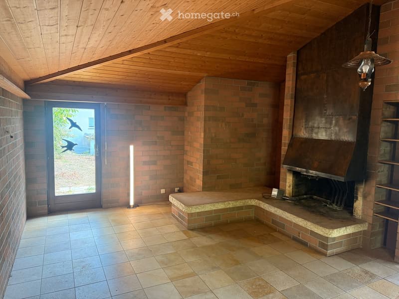 Living room with exposed wooden ceiling, brick walls, fireplace, and large window offering a view of the outdoors.