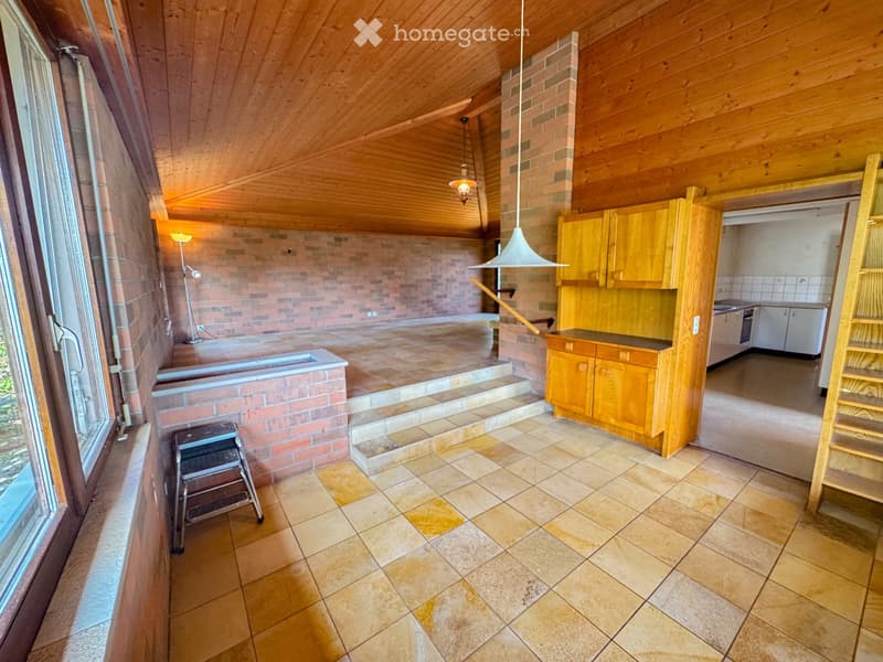 A spacious, well-lit room with wooden beams, brick walls, and tiled flooring. Features include a wooden cabinet, a sink, an oven, a stool, and pendant lights.