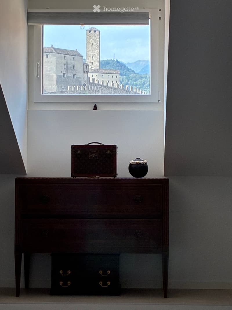 A small wooden chest of drawers with a suitcase and a decorative vase on top, placed against a wall with a large window showing a scenic view of a historic tower and landscape.