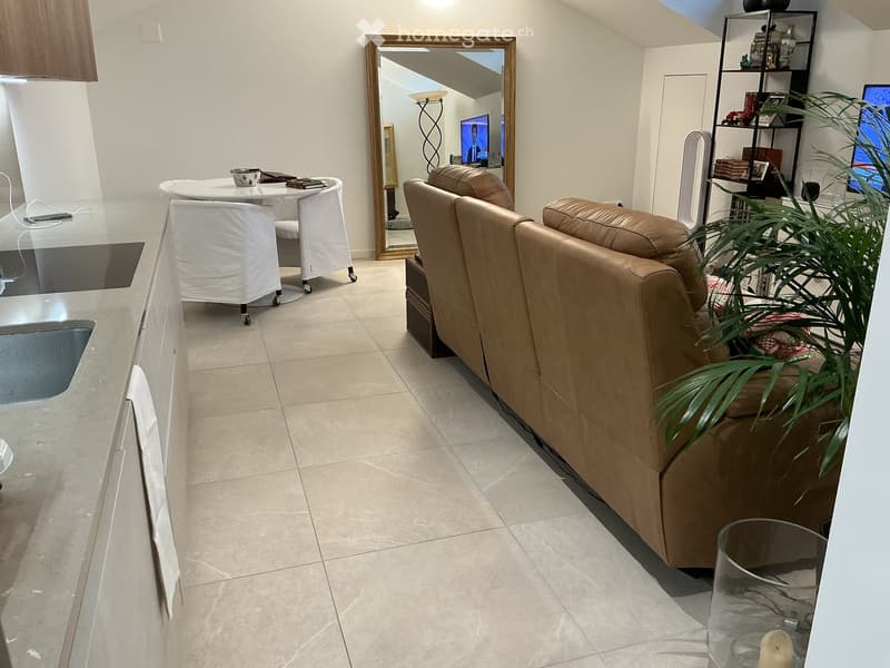 Modern living room with a beige leather sofa, a round white table with chairs, a large mirror, a bookshelf, and a potted plant. The room features tiled flooring and a sleek kitchen area with a glass countertop.