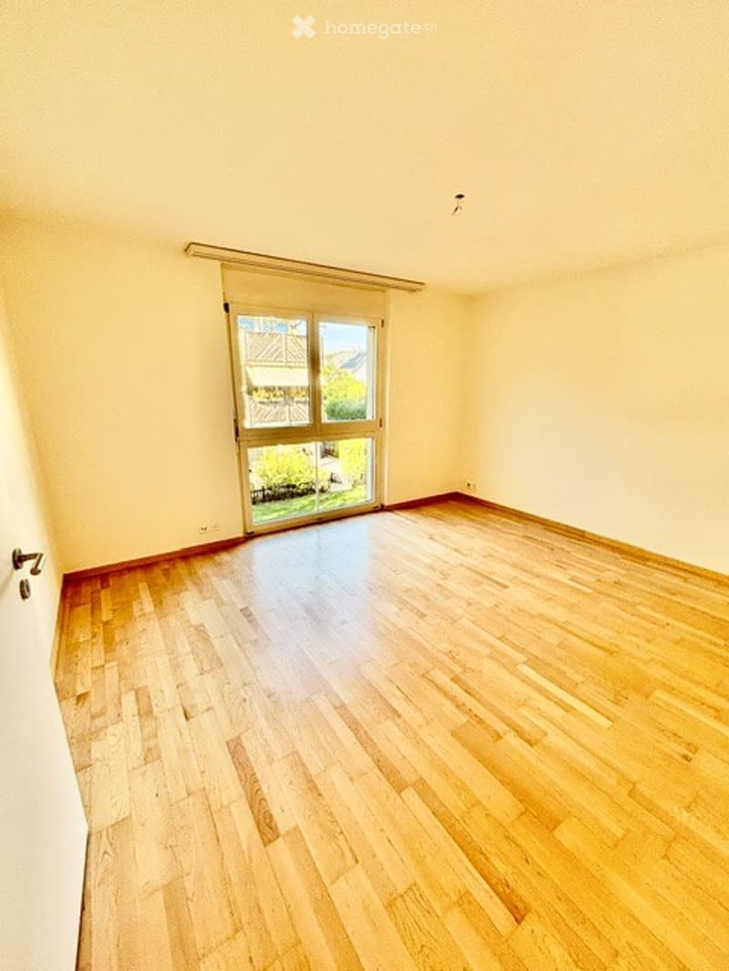 Completely empty room with wooden flooring and large window with blinds, showing a view of the outdoors.